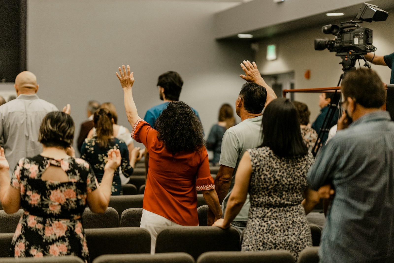 A congregation in worship seen from behind, hands raised, a camera operator filming the service