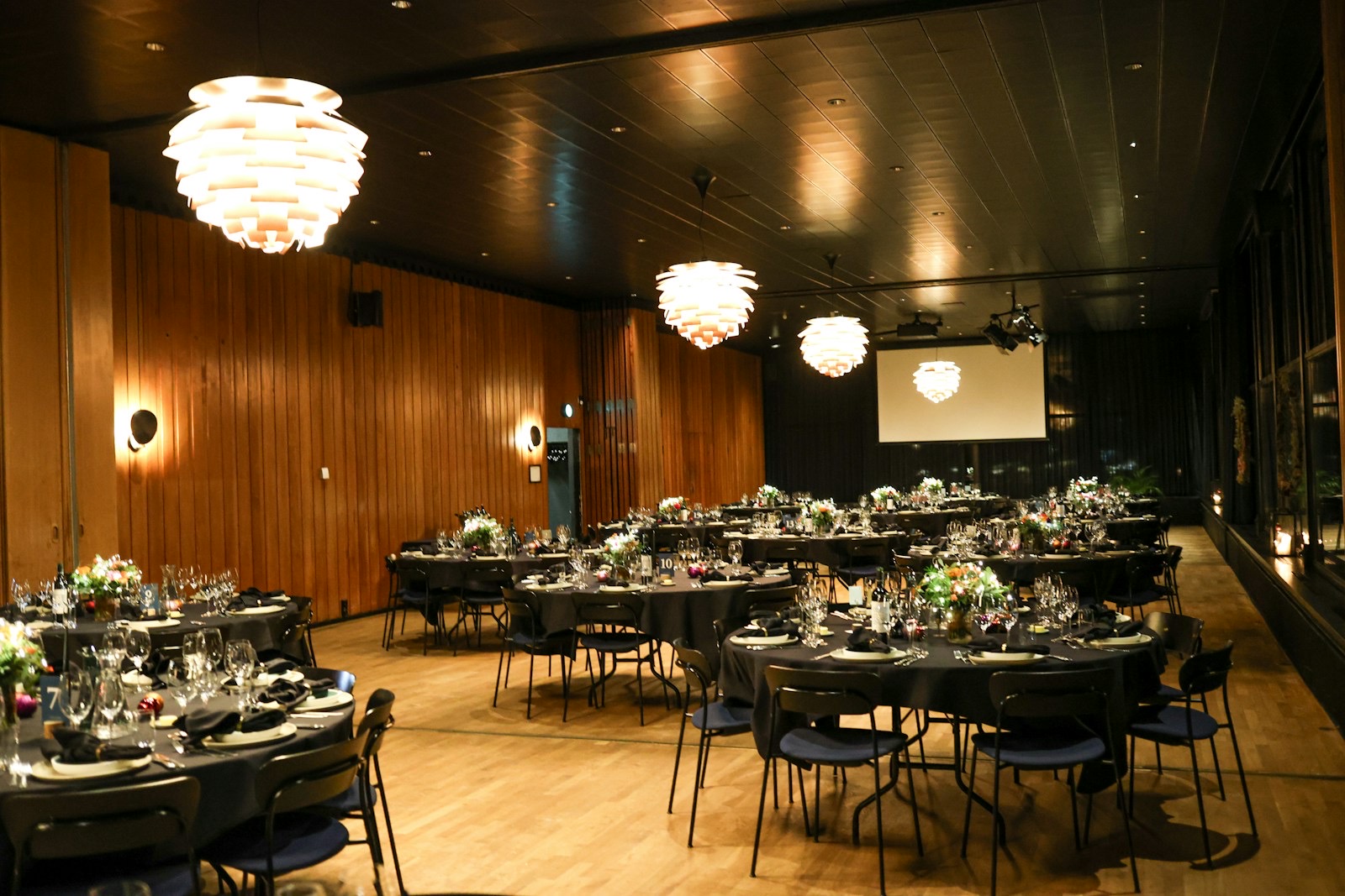 Um local de eventos com mesas redondas sob luminárias pendentes e uma tela de projeção ao fundo