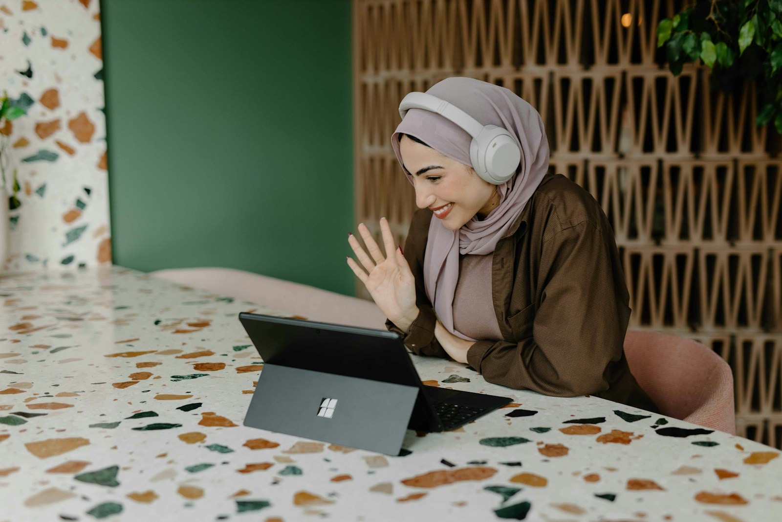 A woman in a headscarf and headphones on a video call at a table in a colourful café