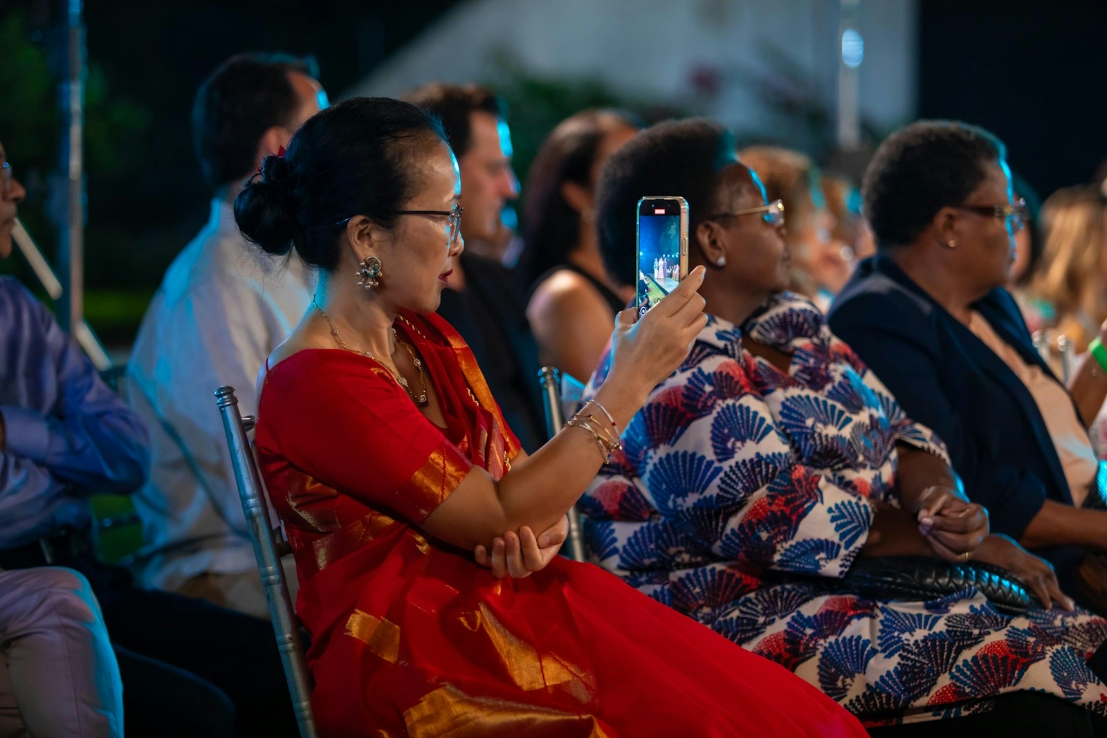 A diverse audience at an evening event; a woman in a red sari holds up her phone